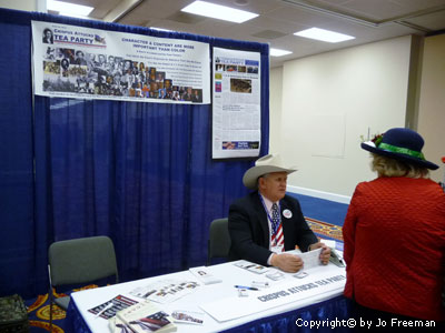 An elderly white man in a cowboy hat and american flag tie sits at a Tea Party booth