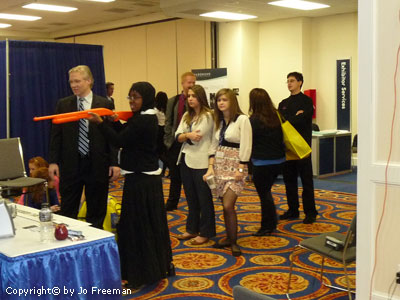 A young african american woman wearing a Hijab and conservative dress aims a bright orage plastic shotgun while other teenagers wait in line