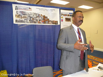 An African American man in a suit gestures at a Tea Party booth